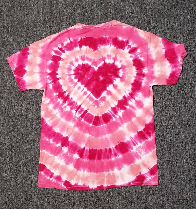 Pu&ograve; includere: Una t-shirt tie-dye rosa e bianca con un design a forma di cuore al centro.