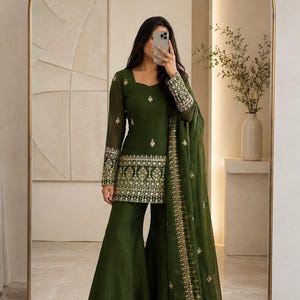 May include: A dark olive green ensemble featuring a long-sleeved tunic with a square neckline and intricate gold embroidery along the hem and cuffs, paired with matching wide-leg trousers and a sheer dupatta with a gold embroidered border.