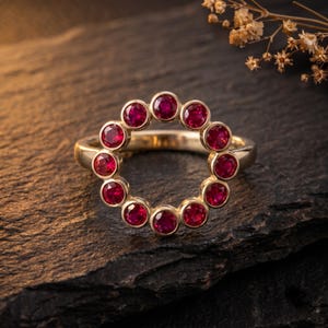 Op de afbeelding: Een gouden ring met een cirkelvormige opstelling van elf ronde, levendig rode robijnen in individuele zettingen. De stenen vormen een doorlopend open cirkelontwerp, waarbij de gepolijste gouden band zichtbaar is achter de cirkelvormige zetting.