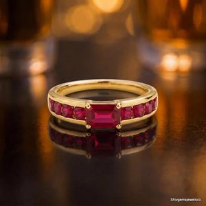 Puede incluir: Un anillo de oro con un rubí rojo central de corte esmeralda, flanqueado por una hilera de rubíes rojos más pequeños de corte redondo engastados en la banda. La joya tiene un acabado en oro amarillo pulido y lleva el texto Shivgemsjewelsco.