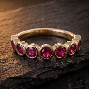 May include: A gold band ring featuring a row of seven round, bezel-set vibrant red rubies. The metal has a polished finish, and the stones are arranged in a continuous line across the front half of the circular band, showcasing a classic design.