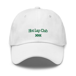 May include: A white baseball cap featuring the embroidered text Hot Lap Club in dark green thread on the front centre. Below the text is a small, dark green embroidered graphic consisting of four connected squares arranged in a horizontal row.