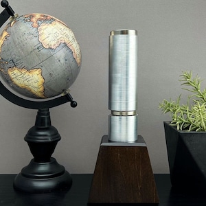 May include: A silver metal cylinder with a wooden base sits on a black surface. A globe and a potted plant are in the background.