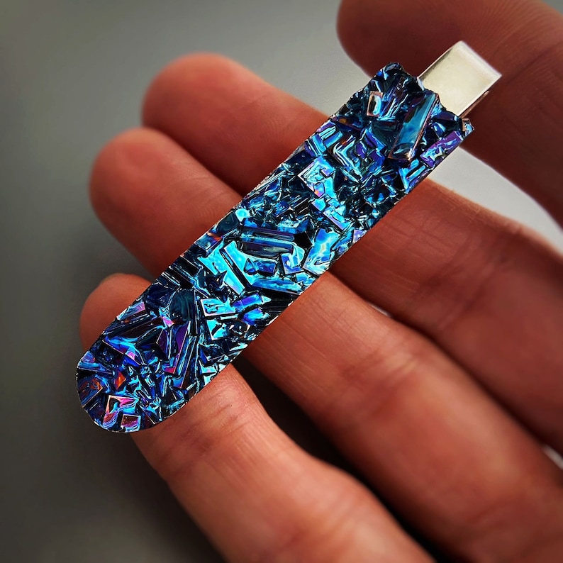 K&ouml;nnte beinhalten: Ein Krawattenclip mit einem blauen, schillernden Kristall. Der Kristall besteht aus Wismut und hat ein einzigartiges, geometrisches Muster.