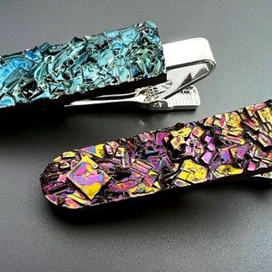 K&ouml;nnte beinhalten: Zwei Krawattenklammern mit einem silbernen Metallclip und einer bunten, schillernden Oberfl&auml;che. Die obere Klammer hat eine blaue und gr&uuml;ne Oberfl&auml;che, w&auml;hrend die untere Klammer eine violette, gelbe und rosafarbene Oberfl&auml;che hat.