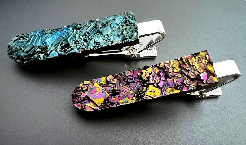 K&ouml;nnte beinhalten: Zwei Krawattenklammern mit einem silbernen Metallclip und einer bunten, schillernden Oberfl&auml;che. Die obere Klammer hat eine blaue und gr&uuml;ne Oberfl&auml;che, w&auml;hrend die untere Klammer eine violette, gelbe und rosafarbene Oberfl&auml;che hat.