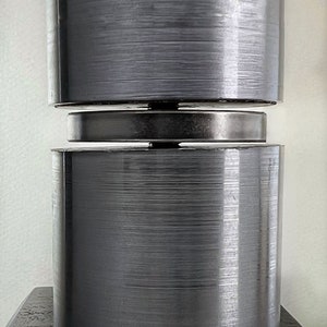 May include: A close-up of a silver metal cylinder with a brushed finish. The cylinder is made up of two stacked pieces, with a smaller, flat, silver metal disc in between.