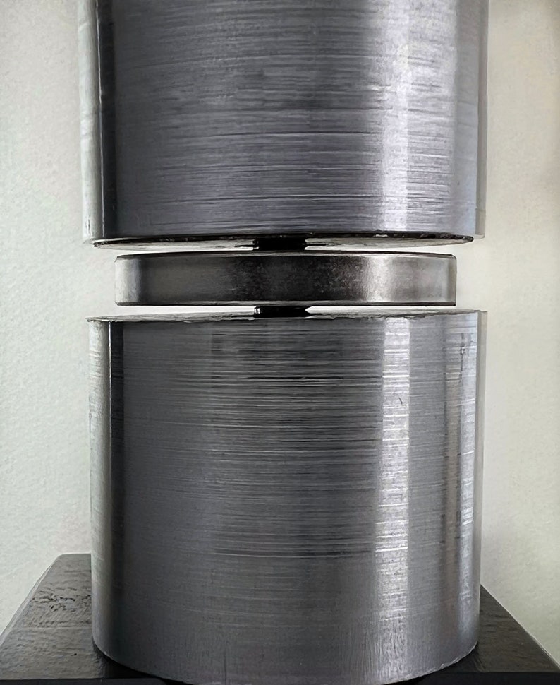 May include: A close-up of a silver metal cylinder with a brushed finish. The cylinder is made up of two stacked pieces, with a smaller, flat, silver metal disc in between.