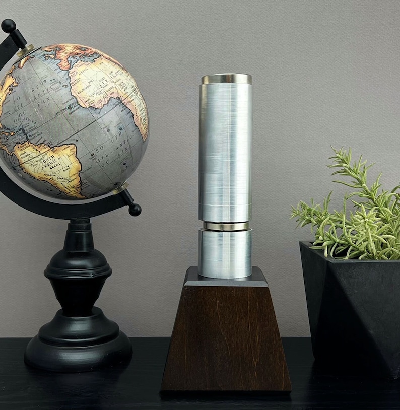 May include: A silver metal cylinder with a wooden base sits on a black surface. A globe and a potted plant are in the background.