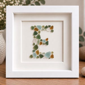 May include: A square white frame containing a piece of wall art featuring the letter E. The letter is constructed from small, smooth pieces of sea glass in various shades of green, brown, and light blue, arranged on a plain white background.