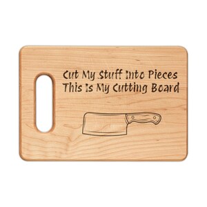 May include: A rectangular light brown wooden cutting board with a handle cutout on the left side. The board features a laser-engraved illustration of a meat cleaver and the text: Cut My Stuff Into Pieces This Is My Cutting Board.