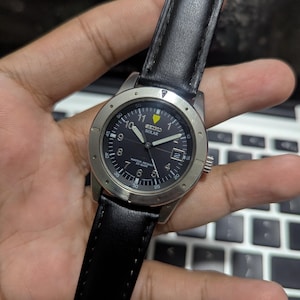 May include: A Seiko Solar wristwatch with a black dial, white Arabic numerals, a yellow triangle marker at the twelve o'clock position, a date window at three o'clock, a silver-tone metal bezel, and a black leather strap with visible stitching.