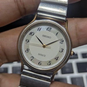 Pode incluir: Um relógio de pulso Seiko Dolce com mostrador redondo na cor creme, algarismos arábicos pretos, ponteiros pretos finos e uma moldura dourada. O relógio possui uma pulseira extensível de metal prateado e uma pequena coroa na lateral.