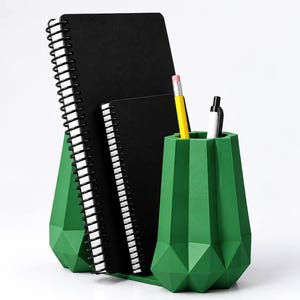 Pode incluir: Um organizador de secretária geométrico verde com superfície facetada, contendo dois cadernos de espiral pretos, um lápis amarelo e uma caneta preta e branca. O texto "Geometric Notebook Holder" está impresso abaixo do objeto.