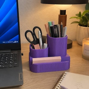May include: A purple desk organizer with a ribbed texture, featuring two cylindrical compartments of varying heights for pens and scissors, and a rectangular front slot for sticky notes. The organizer holds various pens, a pair of black scissors, and a pink eraser.