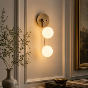 May include: A wall-mounted light fitting featuring a circular brass backplate and a vertical rod supporting two spherical white glass globes. The globes emit a warm, soft glow, and the metal components have a brushed brass finish, creating a balanced, linear design mounted on a wall.