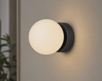 May include: A wall-mounted light fitting featuring a spherical, frosted white glass globe attached to a circular, matte black metal base. The globe emits a soft, warm, diffused glow, illuminating the surrounding area with a gentle light.
