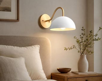 May include: A wall-mounted lamp featuring a matte white dome-shaped shade and a curved brass-toned arm. The fixture is attached to a circular brass-toned wall plate and emits a warm light, positioned above a bed with light-coloured bedding.