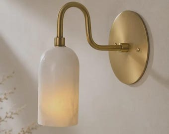 May include: A wall sconce featuring a cylindrical white marble shade with a soft internal glow, attached to a curved brass arm and a circular brass wall plate. The metal components have a brushed finish, and the light emits a warm illumination.