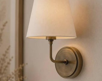 May include: A wall-mounted lamp featuring a circular antique brass backplate and a curved arm supporting a conical off-white fabric lampshade. The fixture emits a soft glow from beneath the shade, highlighting the metallic finish and texture.
