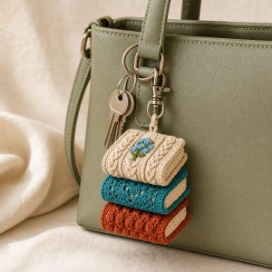 May include: A sage green handbag with a metal keyring attachment featuring three stacked, miniature crocheted books. The top book is cream with blue floral embroidery, the middle is teal with a lace pattern, and the bottom is rust orange.