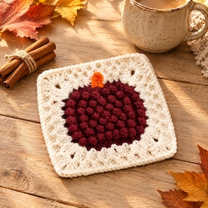 May include: A square crochet coaster featuring a central dark red pumpkin motif with a small orange stem, surrounded by a cream-colored border. The textured design is created with bobble stitches, forming a dense, raised pattern on the surface.