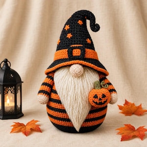 Peut inclure: Une figurine de gnome au crochet portant un chapeau pointu noir avec des étoiles orange et une bande orange. Le gnome a une longue barbe couleur crème, un nez rond et un corps à rayures horizontales noires et orange. Il tient une citrouille.