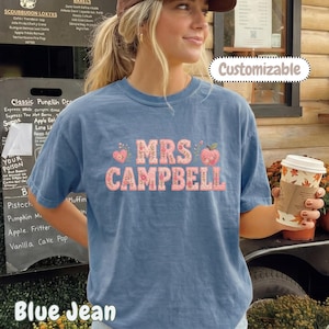 May include: A blue jean coloured short-sleeve t-shirt featuring the text MRS CAMPBELL in a pink floral pattern. The design includes small pink hearts and a floral apple graphic. The shirt has a crew neckline and a relaxed fit, showcasing a casual style.