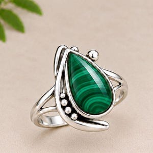 Puede incluir: Un anillo de plata con una piedra preciosa de malaquita en forma de lágrima con bandas verdes distintivas. El engaste de metal incluye un diseño de banda dividida y pequeños acentos esféricos a lo largo del lado de la piedra, con acabado pulido.