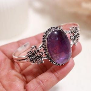 May include: A silver cuff bracelet featuring a large, oval-shaped purple gemstone set in a decorative frame. The band is accented with detailed butterfly motifs on both sides of the central stone, showcasing a polished metallic finish throughout.