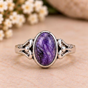 Può includere: Un anello in argento con una pietra preziosa ovale in charoite viola lucida, caratterizzata da inclusioni vorticose nere e bianche. La fascia in metallo presenta un motivo decorativo traforato su entrambi i lati della montatura centrale.