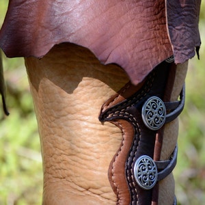 May include: Close-up of a brown leather boot with decorative silver buckles. The boot has a flap that folds over the top and is secured with a buckle.