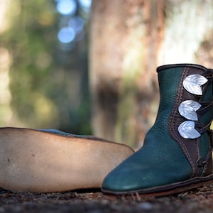 May include: A pair of dark green leather boots with silver leaf accents. The boots have a brown leather trim and laces. One boot is lying on its side, showing the sole. The boots are on a bed of pine needles.