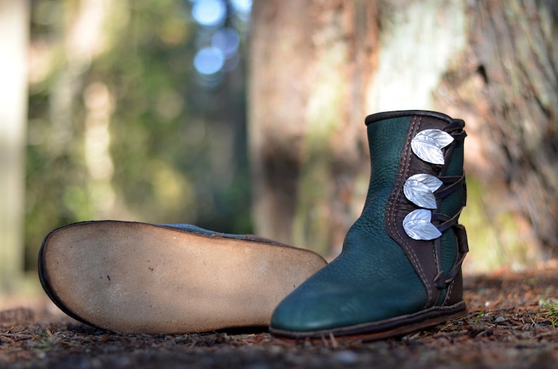 May include: A pair of dark green leather boots with silver leaf accents. The boots have a brown leather trim and laces. One boot is lying on its side, showing the sole. The boots are on a bed of pine needles.