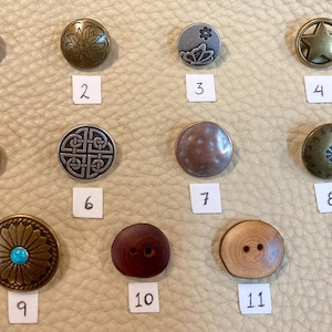 May include: Twelve different metal and wood buttons with various designs, including a star, a flower, a Celtic knot, and a turquoise stone. The buttons are arranged in rows on a light brown surface.
