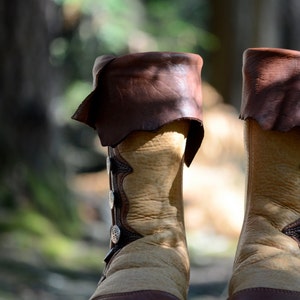 May include: A pair of brown leather boots with decorative silver buckles. The boots are made of a light brown leather and have a dark brown leather trim. The boots are sitting on a bed of brown leaves.