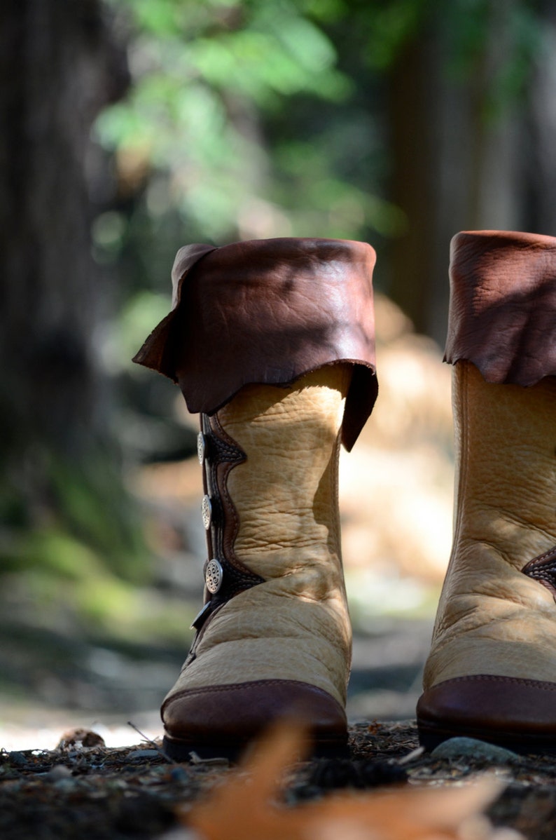 May include: A pair of brown leather boots with decorative silver buckles. The boots are made of a light brown leather and have a dark brown leather trim. The boots are sitting on a bed of brown leaves.
