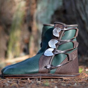 May include: Green leather boots with brown leather accents and silver leaf embellishments. The boots are laced up the front and have a rounded toe.