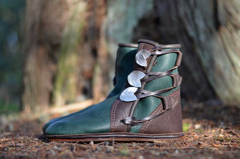 May include: Green leather boots with brown leather accents and silver leaf embellishments. The boots are laced up the front and have a rounded toe.