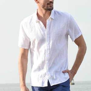 May include: A crisp white short-sleeved button-down shirt featuring a classic collar, a front button placket, and a textured fabric finish. The shirt is worn untucked over dark blue trousers, showcasing a clean, simple, and functional design.