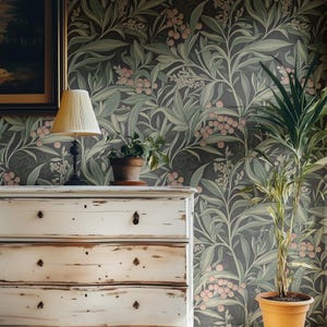 May include: A white dresser with three drawers and black knobs. The dresser is in front of a wall with a dark green floral wallpaper featuring pink flowers and leaves.