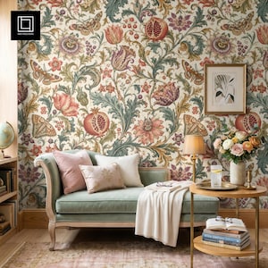 Puede incluir: Una habitación con papel pintado floral con granadas, mariposas y flores en tonos rojos, rosas, verdes y morados. Un sofá verde con cojines rosas y blancos se encuentra frente al papel pintado. Una pequeña mesa con una lámpara y flores.