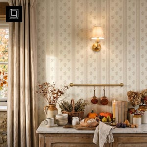 Op de afbeelding: Een keukenscène met crèmekleurig behang met een verticaal bloemenstreepmotief, een messing wandlamp met stoffen kap, een messing rail met twee koperen pannen, een keramische kan, een gevlochten mand en een marmeren aanrechtblad.