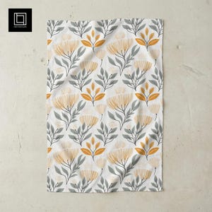 May include: White floral patterned kitchen towel. The design features stylized yellow flowers, gray leaves, and orange accents. The towel is made of a soft, absorbent fabric, perfect for drying dishes or wiping surfaces.