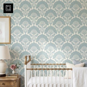 Pode incluir: Um padrão de papel de parede floral azul claro e creme. O design apresenta formas de flores estilizadas dispostas em um padrão repetitivo. Um berço branco com detalhes dourados e uma mesa de cabeceira de madeira com uma lâmpada e um vaso de flores estão em primeiro plano.
