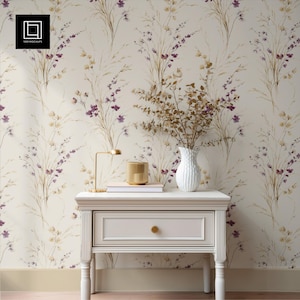 May include: A white nightstand with a drawer, a gold lamp, and a gold candle holder. A white vase with dried flowers sits on top of the nightstand. The wallpaper has a floral pattern with purple and beige accents.