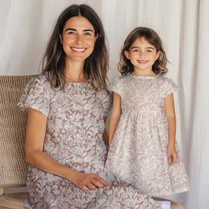Può includere: Una madre e una figlia che indossano abiti a fiori abbinati. Gli abiti hanno uno sfondo rosa chiaro con un motivo floreale bianco. La madre indossa un abito a maniche corte e la figlia indossa un abito a maniche corte con una gonna leggermente svasata.