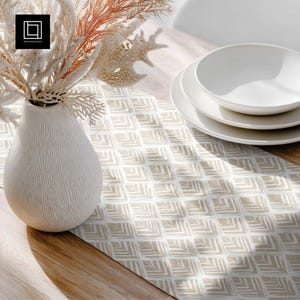 May include: A white ceramic vase with a textured surface holds dried flowers and coral. A stack of white plates and bowls sits on a beige and white patterned table runner. The scene is set on a wooden table.