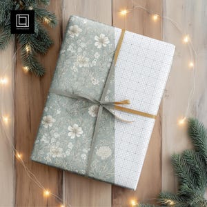 Puede incluir: Un regalo envuelto con dos papeles diferentes. Uno presenta un patrón floral en tonos verdes y blancos, el otro un patrón de cuadrícula. El regalo está atado con una cinta gris y una cinta dorada. Luces parpadeantes y ramas de pino rodean el regalo.