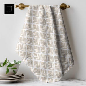 May include: A white and beige geometric patterned tea towel hanging from a brass rail. The tea towel features a repeating square design with angled lines in a neutral colour. The tea towel is displayed in a kitchen setting.